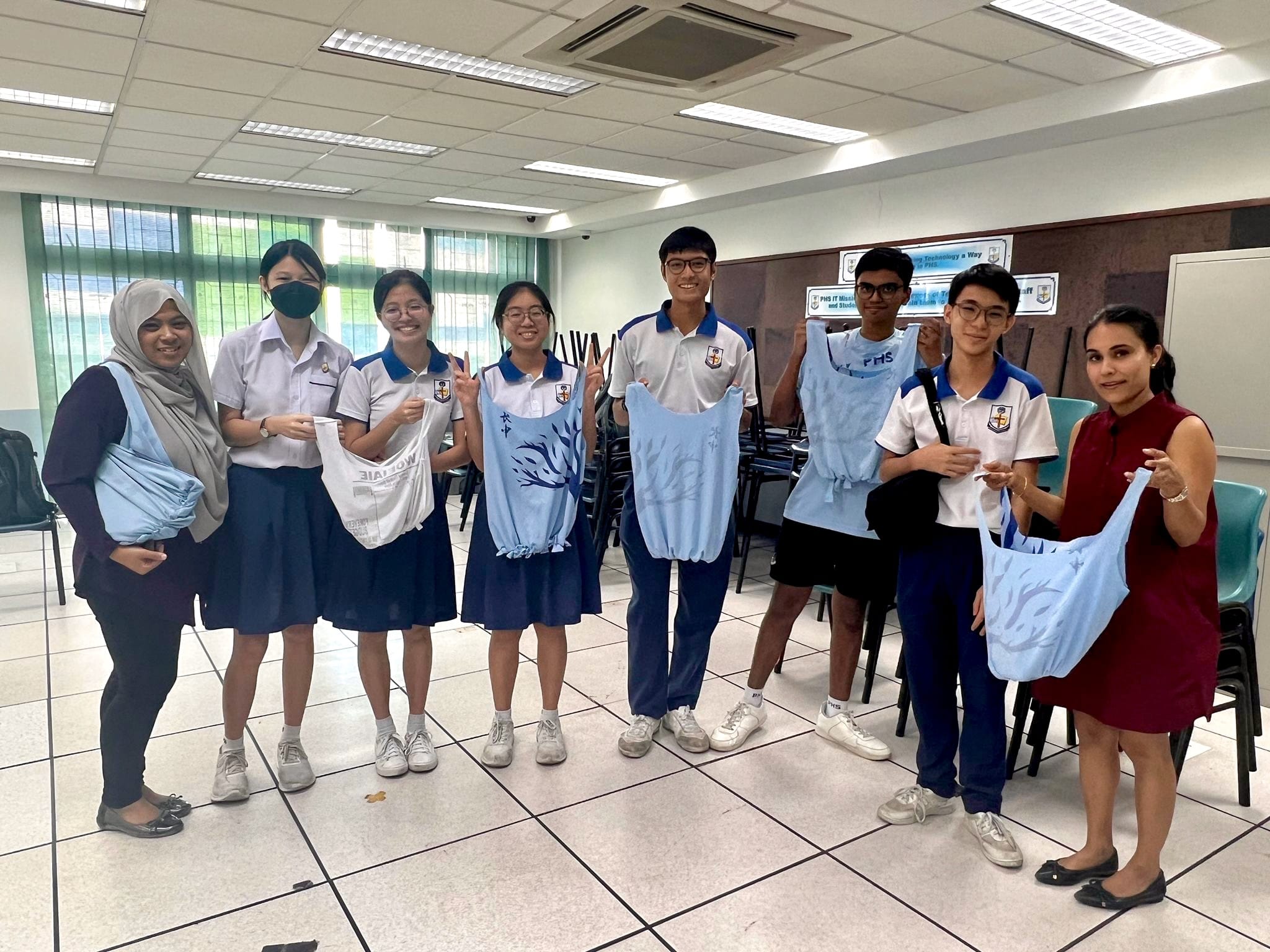 Six students in school uniforms and two adults stand in a classroom holding light blue tote bags.